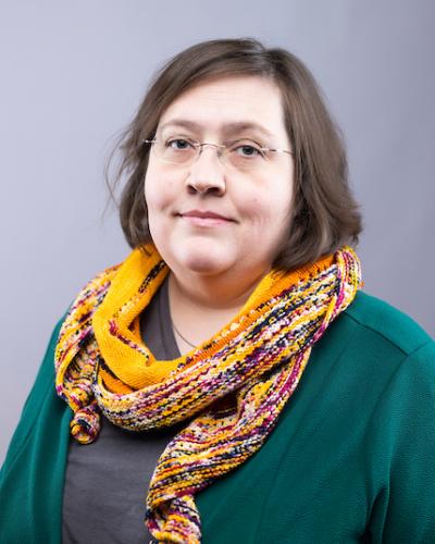 A woman with brown hair, wearing glasses, a green jacket, and a yellow and multicolored scarf.