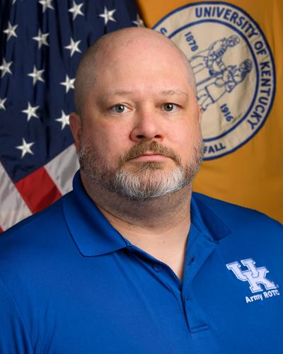 A man wearing a blue University of Kentucky polo shirt stands in front of an American flag and University of Kentucky seal.