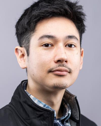 A man wearing a black jacket and blue plaid shirt looks at the camera against a gray background.