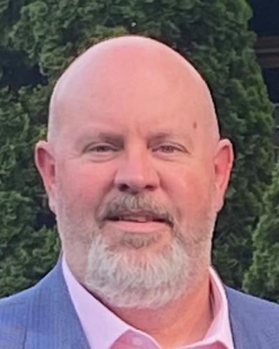 A man in a blue suit jacket and pink collared shirt smiles while standing in front of several trees.