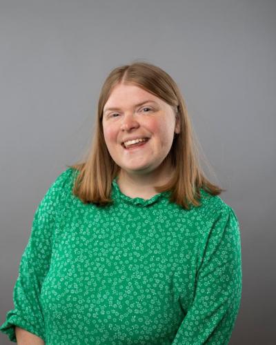 A woman with shoulder-length hair wearing a green patterned blouse smiles while posing in front of a gray background.