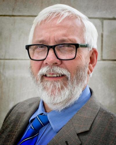 A man with white hair, wearing black glasses, a blue shirt, tie, and gray suit jacket, smiling while standing in front of a stone wall.