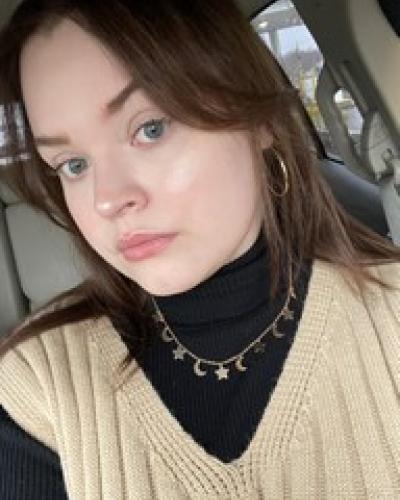 A young woman with brown hair and a neutral expression, wearing a black turtleneck, tan sweater, and jewelry, sits in a car.